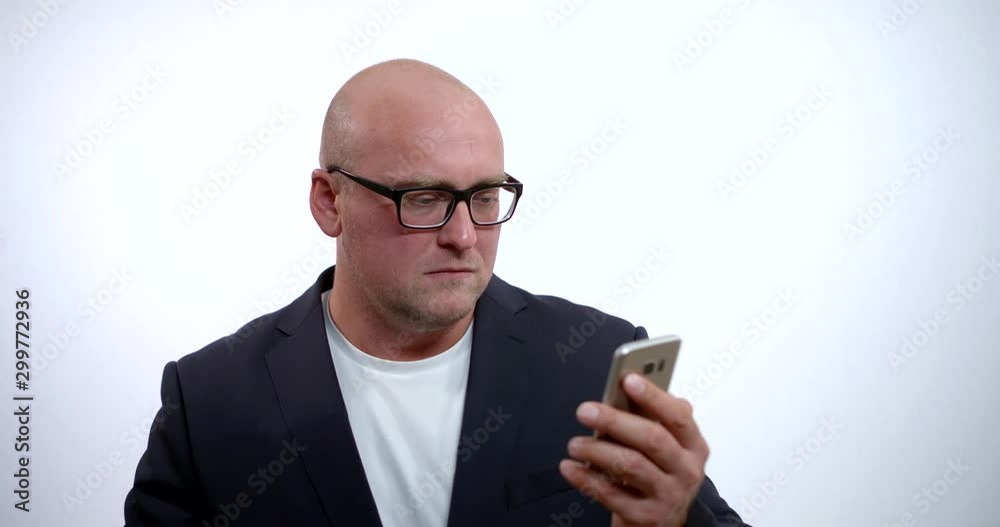 Portrait of a bald middle-aged business man wearing glasses and a black jacket, he is in the Studio on a white background and looking at his mobile phone. Portrait of a bald middle-aged business man wearing glasses and a black jacket, he is in the Studio on a white background and looking at his mobile phone.
