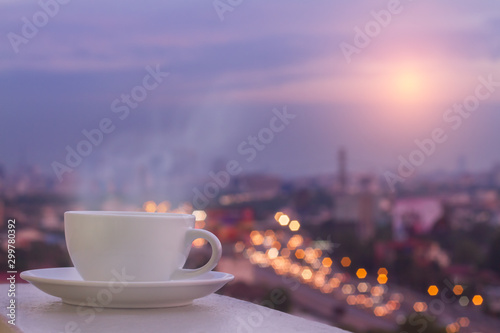 White coffee cup and softy stream smoke put on sky terrace border with blurred sunrise high angle view of city scape and colorful traffic lighting on road. Selective focus and copy space.