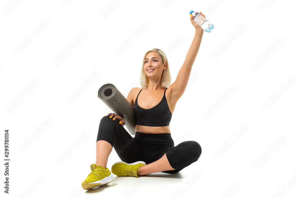 Sport caucasian girl sits on floor, cross legs, keeps a gymnastic mat and bottle of water isolated on white background Sport caucasian girl sits on floor, cross legs, keeps a gymnastic mat and bottle of water isolated on white background