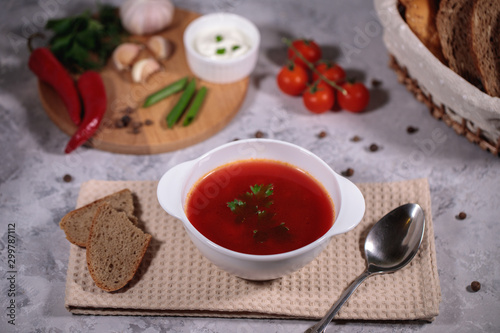 Tasty and hearty dinner. A plate with borsch on the table, next to the board are vegetables, parsley, dill, green onions, garlic, chili peppers, cherry tomatoes, and bread. photo