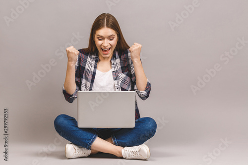 Winner! Excited smiling girl sitting on floor with laptop, raising one hand in the air is she wins, isolated on grey background. © denis_vermenko