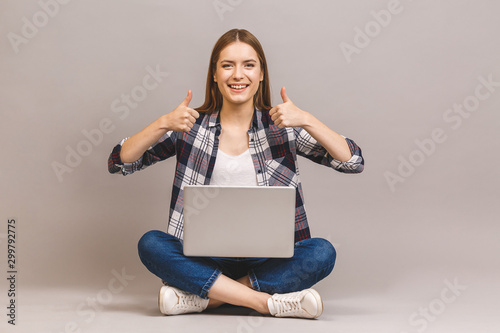 Happy young woman sitting on the floor with crossed legs and using laptop isolated on gray background. Thumbs up. © denis_vermenko