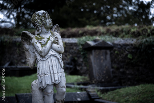 KILKENNY, IRELAND, DECEMBER 23, 2018: Sculpture of an old creepy cherub angel in the middle of a graveyard, full of lichen and mold, holding its chest and heart while looking at the cloudy sky.