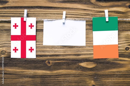 Hanging flags of Georgia and Ireland attached to rope with clothes pins with copy space on white note paper on wooden background.Diplomatic relations between countries. photo