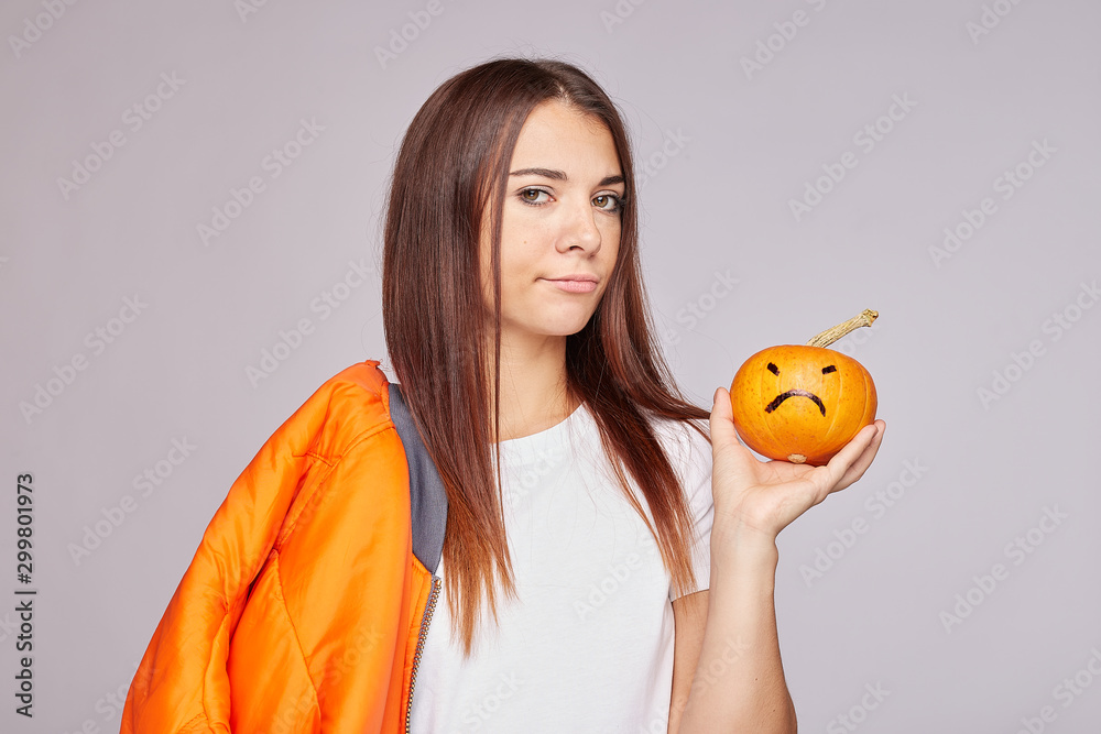 Shot of mixed raced young woman with displeased sad face expression, focuses at camera with pumpkin in hand, wears white casual clothes, stand on grey wall background with copy space for advertisement Shot of mixed raced young woman with displeased sad face expression, focuses at camera with pumpkin in hand, wears white casual clothes, stand on grey wall background with copy space for advertisement