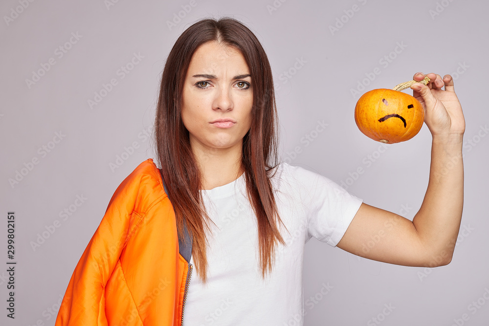 Indoor shot of confused displeased woman, holds very small pumpkin, has puzzed look, smirks face, wears white blank shirt, isolated on grey, feels sad, not ready for holiday. Party time concept. Indoor shot of confused displeased woman, holds very small pumpkin, has puzzed look, smirks face, wears white blank shirt, isolated on grey, feels sad, not ready for holiday. Party time concept.