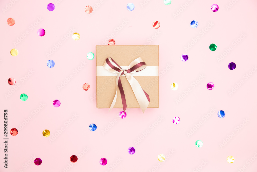 Gift box with kraft cardboard bow on pink background with confetti, top view. Surprise concept, birthday present