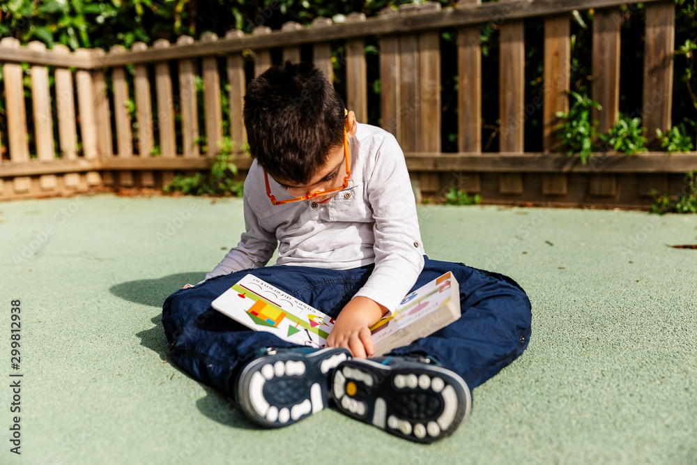 Fototapeta premium Niño con pluri discapacidad leyendo, mirando, jugando con un libro de colores en el suelo de un parque de ciudad.