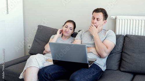 Couple is watching bored film and falling asleep. Young man and woman watch video on laptop sitting on sofa. They are in their modern flat.