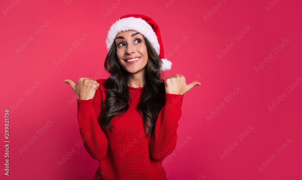 Everything is going swimmingly. A smiley young lady dressed in red for New Year celebration is pointing with thumbs of both of her hands sideways and looking to the right upper corner. Everything is going swimmingly. A smiley young lady dressed in red for New Year celebration is pointing with thumbs of both of her hands sideways and looking to the right upper corner.