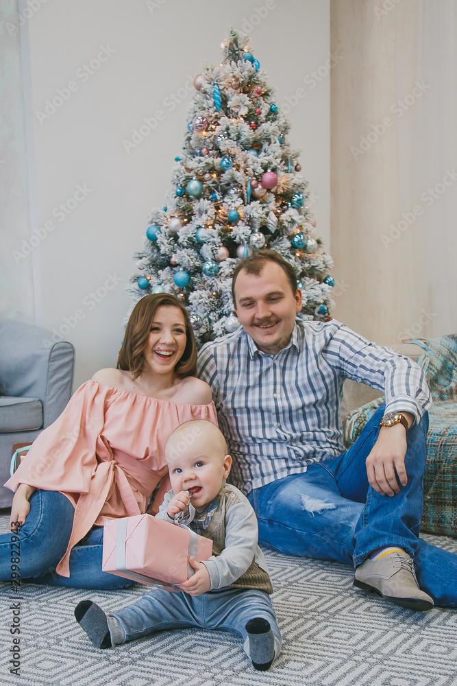 happy family new year photo session: mother, father and little child are sitting near the new year tree in a bright interior 