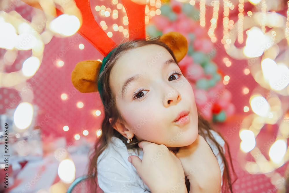 Bokeh lights from a Christmas garland on the forefront and funny little girl with reindeer antlers, looking at the camera, smiling in the evening at home. Bokeh lights from a Christmas garland on the forefront and funny little girl with reindeer antlers, looking at the camera, smiling in the evening at home.