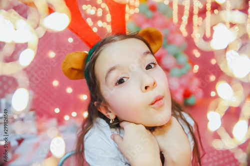 Bokeh lights from a Christmas garland on the forefront and funny little girl with reindeer antlers, looking at the camera, smiling in the evening at home.
