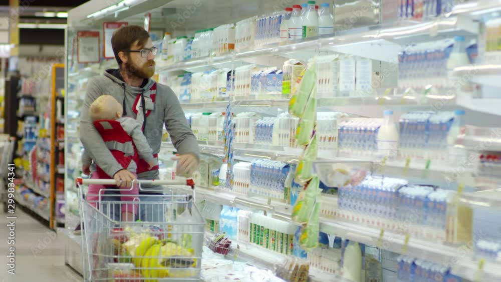 Front view of Caucasian man in glasses carrying baby in sling, talking to child and putting bottle of milk into shopping trolley at supermarket