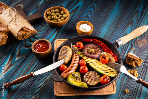 Grilled sausages and vegetables in a pan  towel  mayonnaise  olives and ketchup in wooden bowls  broken buckwheat baguette and cutlery on a blue wooden background.