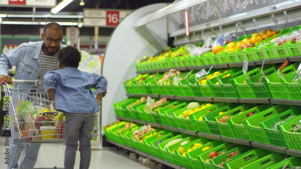 Front view of black man in glasses having fun with little son at supermarket and jumping while pushing shopping cart with goods, boy riding it