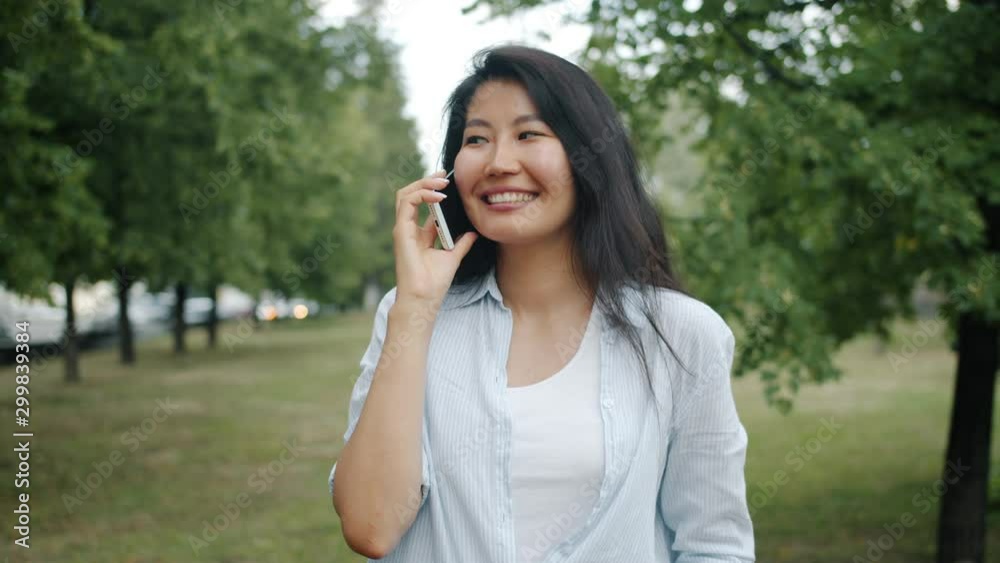 Joyful Asian woman student is chatting on mobile phone in green park in summer enjoying casual conversation outdoors. Youth and modern technology concept. Joyful Asian woman student is chatting on mobile phone in green park in summer enjoying casual conversation outdoors. Youth and modern technology concept.