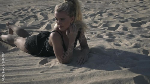 Young beautiful woman with blonde hair and make up in black shine swimsuit lying alone and sunbathing on white sand beach under sunlights.