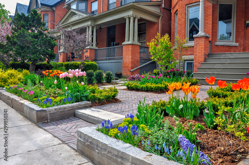This beautiful urban front yard garden features a large veranda, brick paver walkway, retaining wall with plantings of bulbs, shrubs and perennials for colour, texture and winter interest.