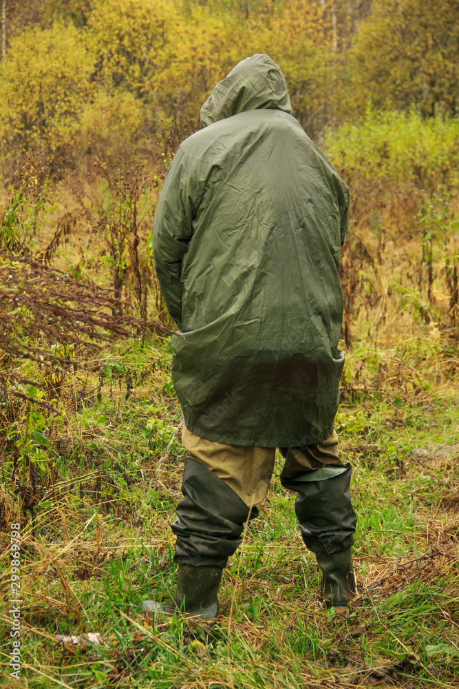 A man in high boots and camouflage stands and urinates in the woods. Rear view. The concept of extreme travel in the taiga.