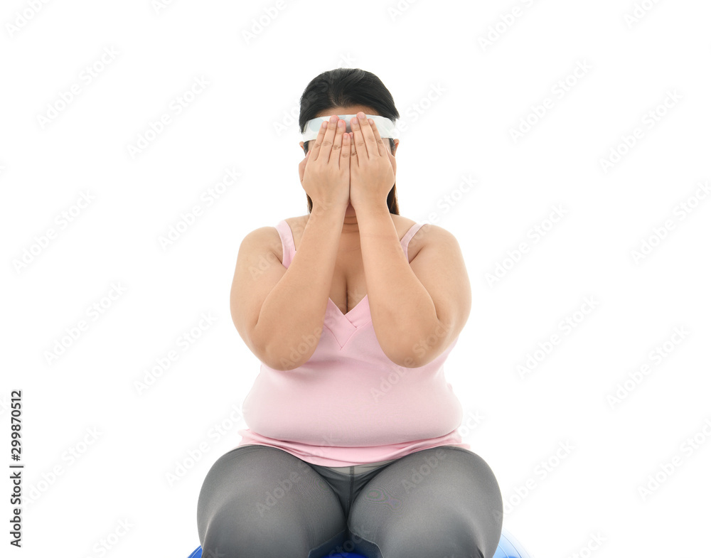 Overweight Asian woman sitting on a gym ball looking stressed covers her face with her hands isolated on white background