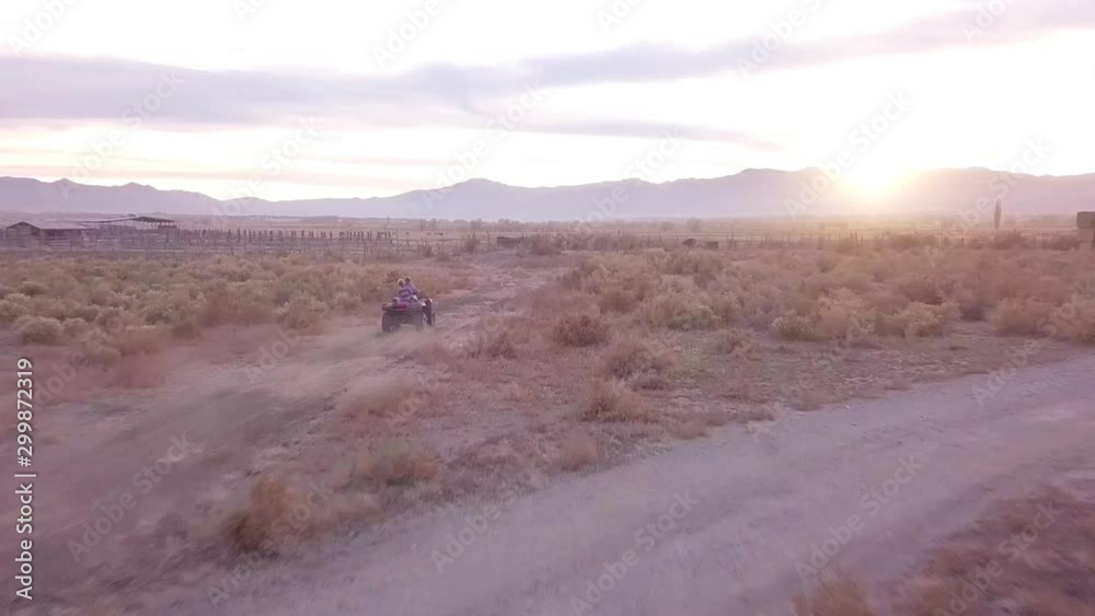 Drone shot following kids riding an ATV (four-wheeler) down a dirt path, creating lots of dust, on their ranch in Utah. Shot during a golden hour with a beautiful sunset on the horizon. Drone shot following kids riding an ATV (four-wheeler) down a dirt path, creating lots of dust, on their ranch in Utah. Shot during a golden hour with a beautiful sunset on the horizon.