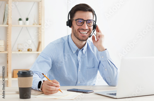 Smiling male customer service worker, sitting with laptop at desk in white modern office