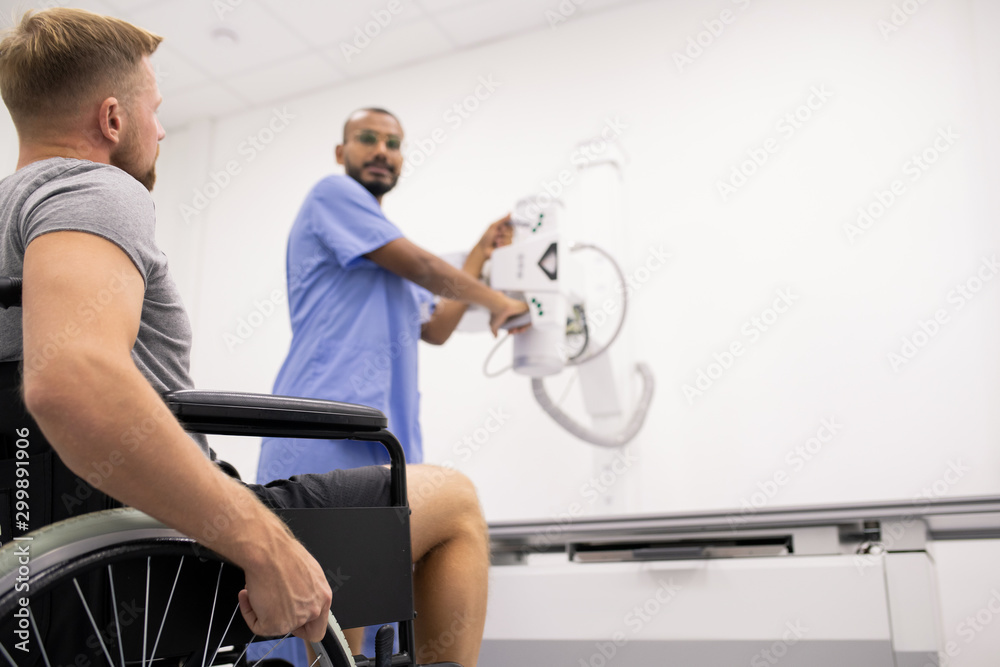 Young man with broken leg sitting on wheelchair and looking at radiologist
