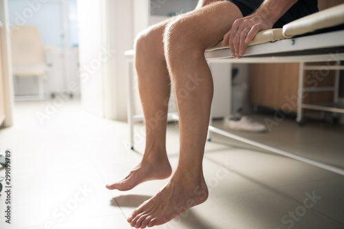 Bare legs of sick male patient sitting on couch in medical office