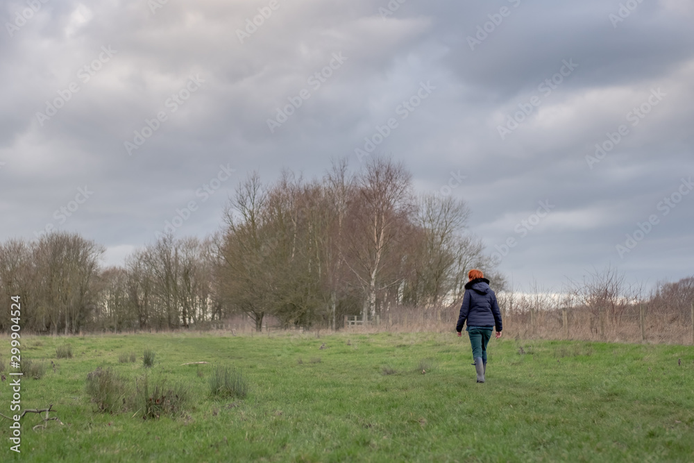 Middle aged woman with auburn hair seen walking away from the camera, along a grassy bank next to an inland river in springtime