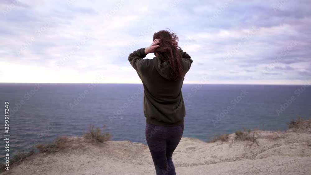A beautiful young woman athlete walks to the edge of a cliff on a windy day. Sports sweatshirt, strong wind disheveles the girl's long hair A beautiful young woman athlete walks to the edge of a cliff on a windy day. Sports sweatshirt, strong wind disheveles the girl's long hair