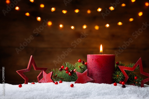Christmas or advent candle, fir branches, berry and red stars in snow against light garland background. Holiday card.