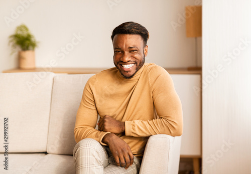 Afro Guy Smiling At Camera Sitting On Sofa At Home © Prostock-studio