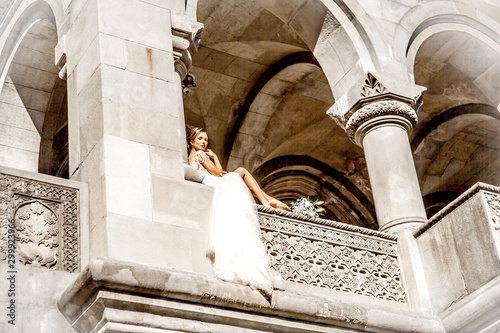 Young beautiful bride wearing elegant wedding dress pozing in old street of the city.  Woman enjoy the sun on the balcony of an old building. photo