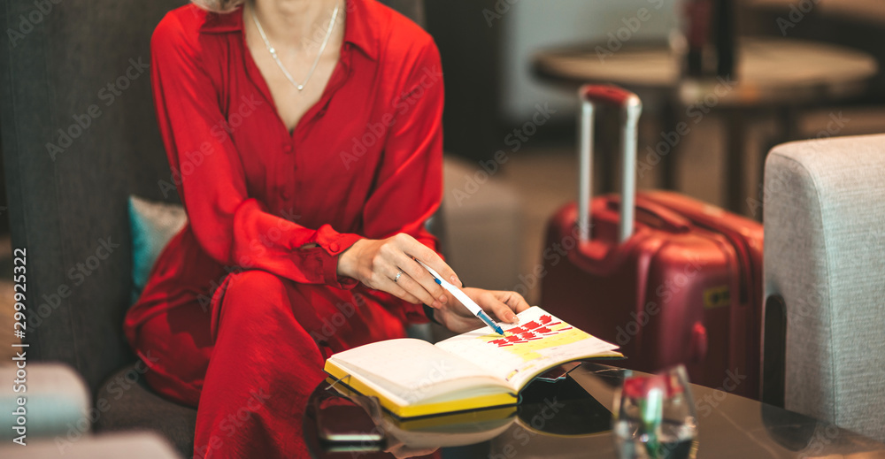 Cropped portrait of gorgeous lady with blonde hair wearing red jumpsuit, looking at notes in her notebook while sitting on the couch. Successful beautiful woman concept. Horizontal shot. Front view - obrazy, fototapety, plakaty 
