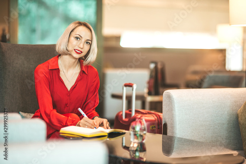 Portrait of gorgeous lady with blonde hair wearing red jumpsuit  looking at camera while making notes in her notebook sitting on the couch. Successful beautiful woman concept. Horizontal shot