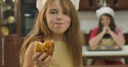 Small Caucasian daughetr looking back at her mom,. turning to camera and chewing sweet baked cookie. Little cute girl eating bun at the kitchen at home. Cinema 4k footage ProRes HQ. photo