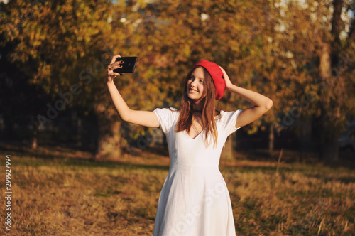 Outdoor shot of lovely girl wearing beret in autumn park makes self portrait on her cellphone. Girl holding smartphone making photo in open air for social networking. People concept