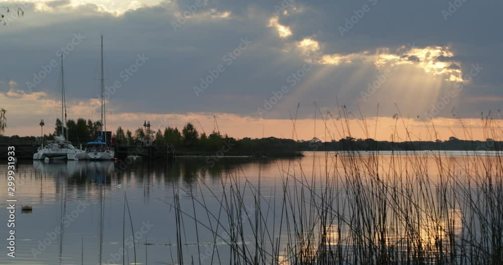 Foreground of wetland plants, sailboats moored at wood dock at background at sunset. Orange reflections over water and volume light. Villa Soriano, Uruguay Foreground of wetland plants, sailboats moored at wood dock at background at sunset. Orange reflections over water and volume light. Villa Soriano, Uruguay