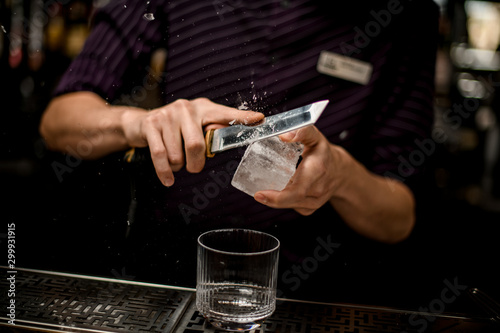 Male bartender cutting ice with a knife above the empty cocktail glass
