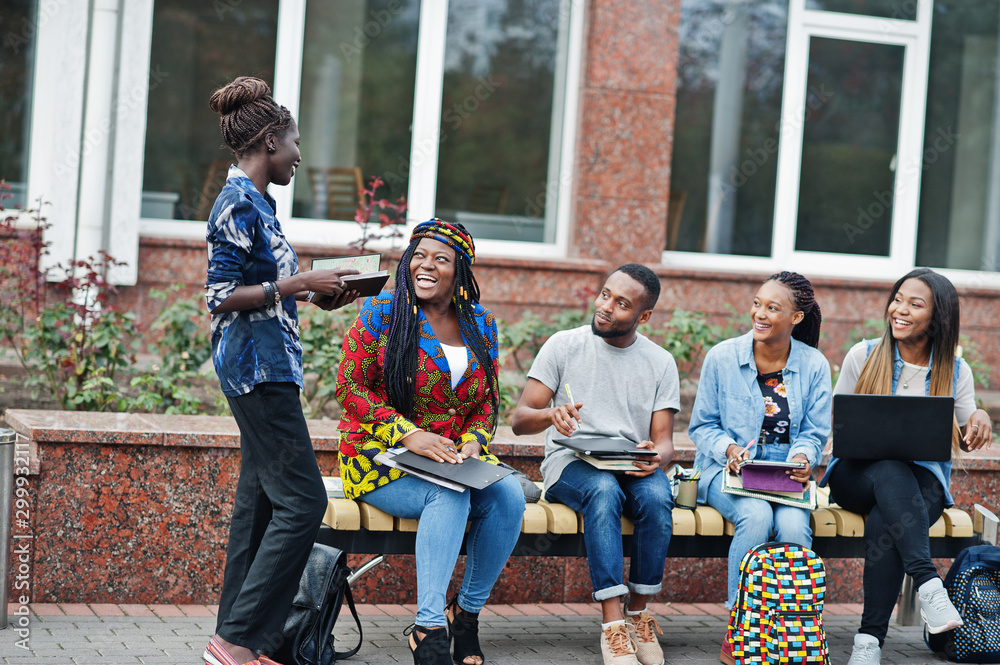 Group of five african college students spending time together on campus at university yard. Black afro friends studying at bench with school items, laptops notebooks.
