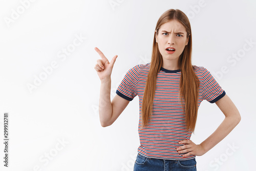 Distressed frustrated, serious-looking young bothered woman with fair long hair, frowning squinting disappointed, pointing upper left corner, demanding explanations, standing white background