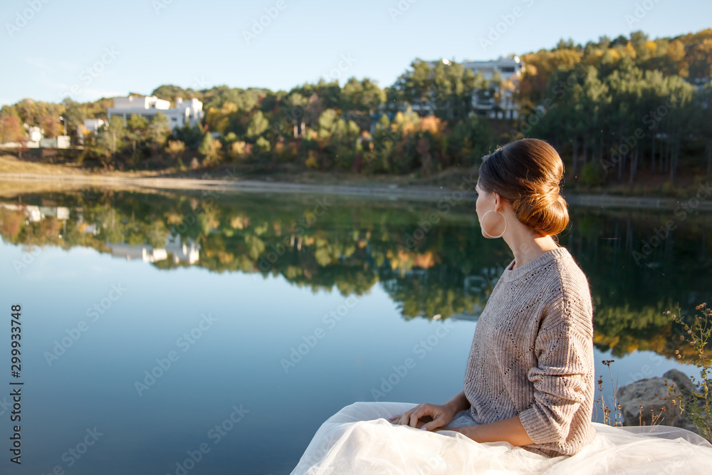 Rear view of young beautiful lonely bride sit on the nature grass and look to the lake and forest