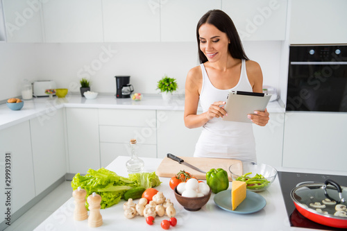 Portrait of beautiful positive brown haired woman want cook tasty yummy healthcare dinner use tablet find recipe follow look at useful fresh ingredients wear white singlet in house kitchen