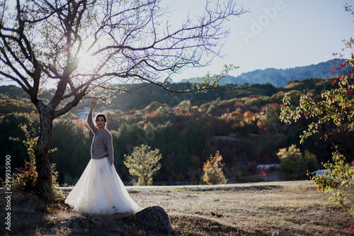 Young beautiful lonely bride posing on the nature and look to the lake and forest. Female wearing wedding dress and sweater at the background trees against the sunset.