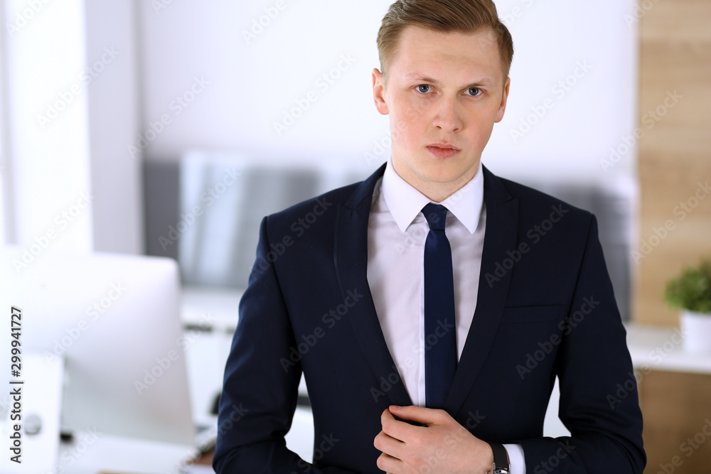 Cheerful businessman standing and looking at camera in office. Headshot of young and successful entrepreneur. Business concept Cheerful businessman standing and looking at camera in office. Headshot of young and successful entrepreneur. Business concept