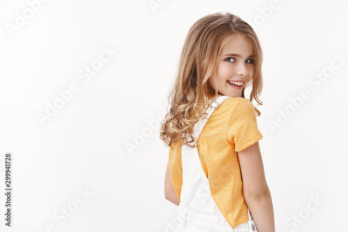 Tender lovely self-assured little girl with blond hair turn behind look camera sly and amused, grinning myserious have something interesting in mind, stand white background wear t-shirt and overalls photo