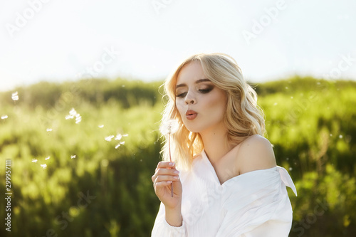 Girl in a long white dress with a dandelion flower in her hand stands in grass in a field. Blonde woman in the sun in a light dress. Girl resting and dreaming  perfect summer makeup on her face