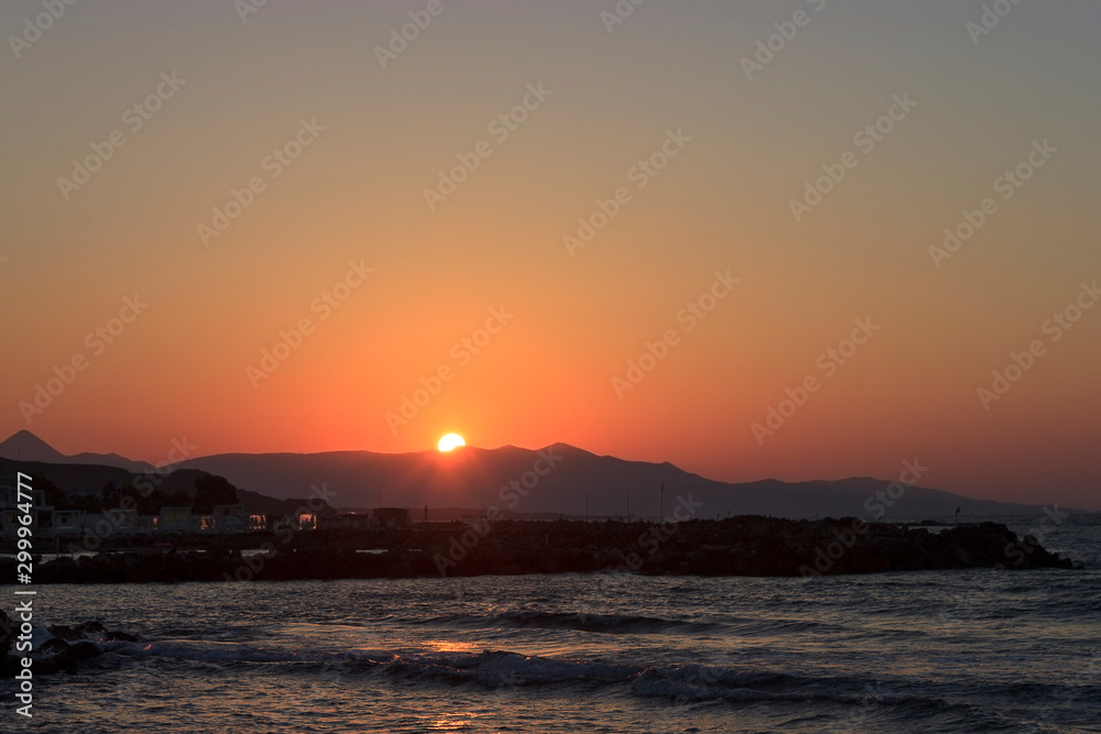 Naklejka premium Sunset over the sea in Kokkini Hani, Crete, Greece. The sun disappears behind the mountain. Scenic seaside landscape in the evening.