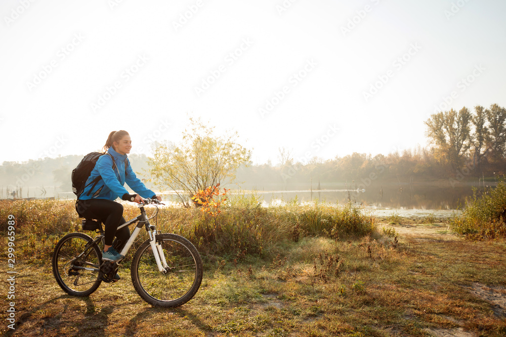 Smiling happy young woman riding mountain bike by the lake or river in early morning sunshine. Sun rising above water in the background. Active and healthy lifestyle.